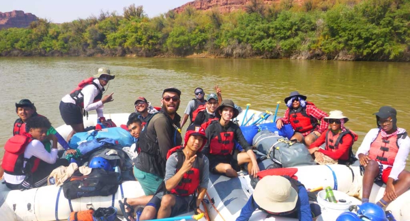 A group of people wearing life jackets smile for the photo as they sit in rafts floating on a calm river. 